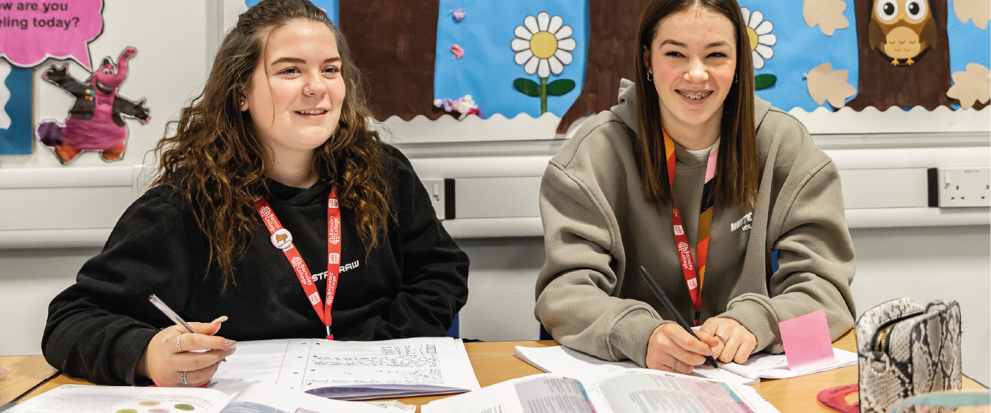 two students smiling with pens in their hand and books in front on them on a desk