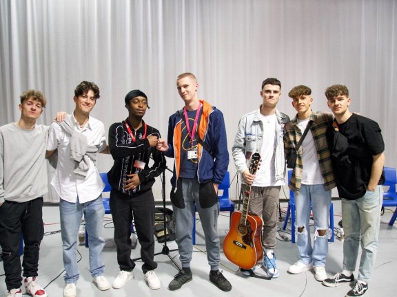 A music group consisting of seven young men. One holds a guitar and two members of the band fist bump.