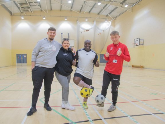 Three students dressed in sports wear pose with a footballer, who is balancing a football on his foot