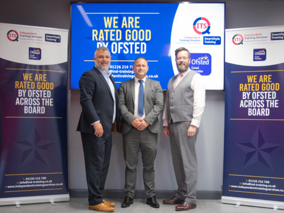 Three people stood in front of a tv screen with two banners stating ITS have achieved good across the board when judged by OFSTED.