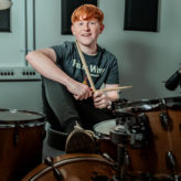 Finley Feast is sat at a drum kit smiling to camera.