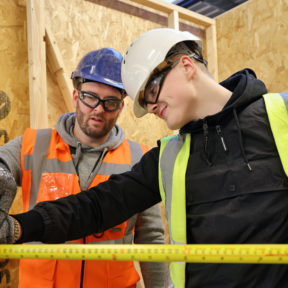 Two people wearing hi-vis jackets, safety hats, goggles and gloves. The person on the right is holding a tape measure.