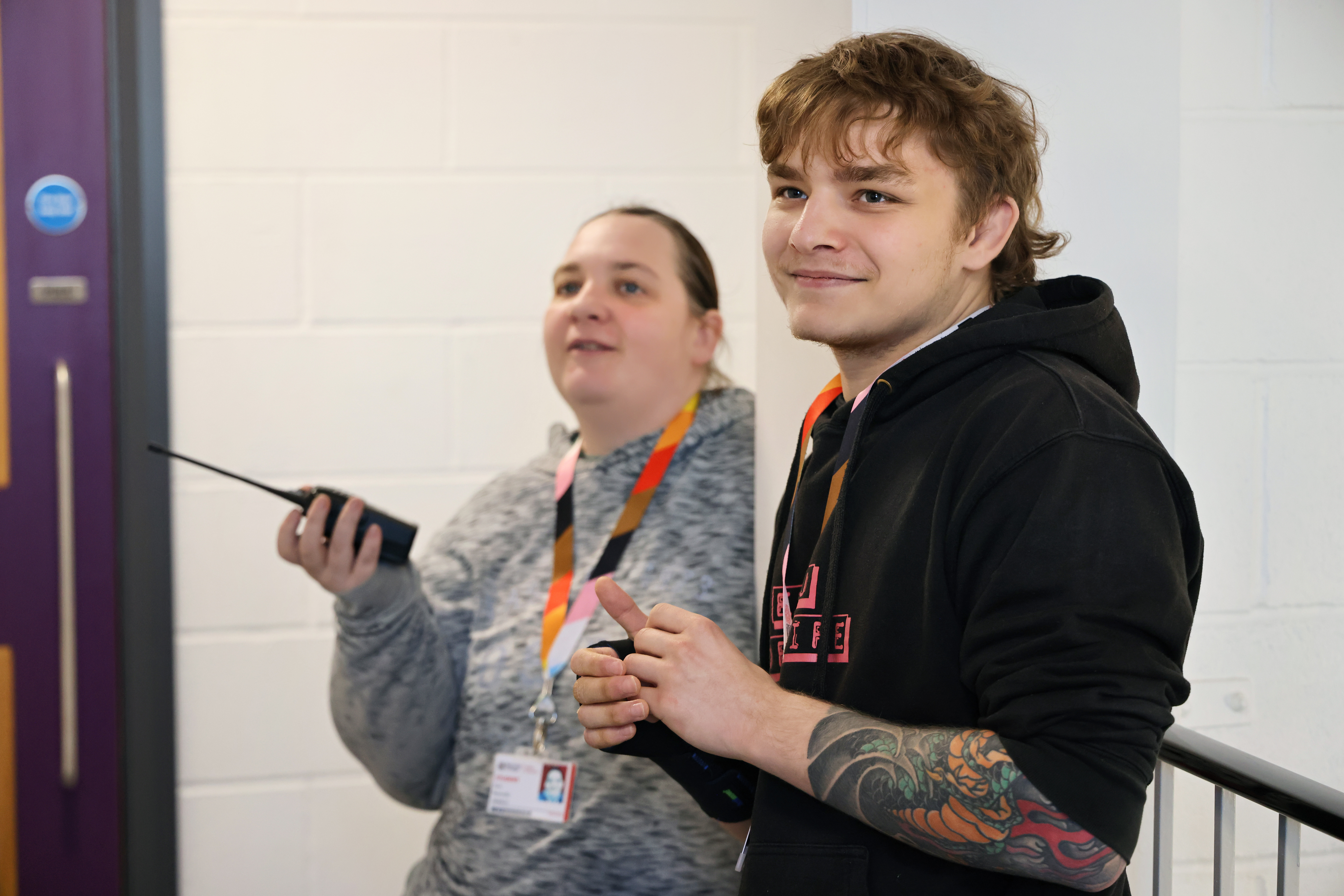 One female and one male are stood side by side. The male is smiling. The female is holding a walkie-talkie.