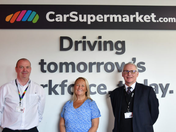 Three people stood in front of a white wall with Car Supermarket logo and the words "Driving tomorrow's workforce today."