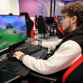 A male is sat in a ref and black gaming chair, looking at a monitor which has a blue car driving though a field on the screen.