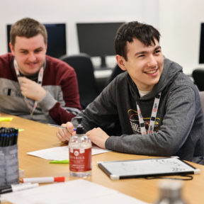 Two students sat at a desk with pens and paper, smiling.