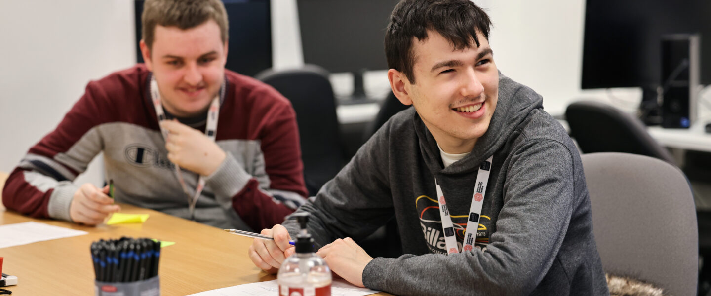 Two students sat at a desk with pens and paper, smiling.