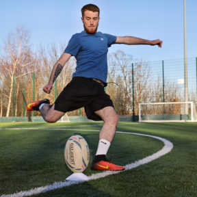 A male wearing a blue top and black shorts, preparing to kick a rugby ball.